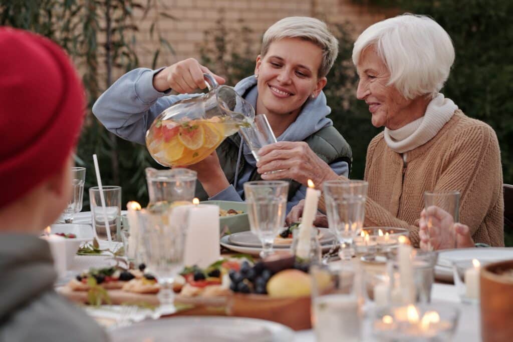 Two older women sit at a table full of food and beverages. One woman pours a pitcher into the other's glass.