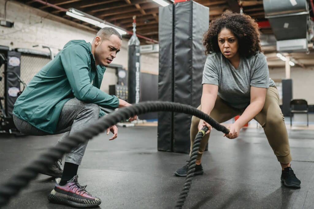 A personal trainer crouches beside his client, who is hard at work using battle ropes.